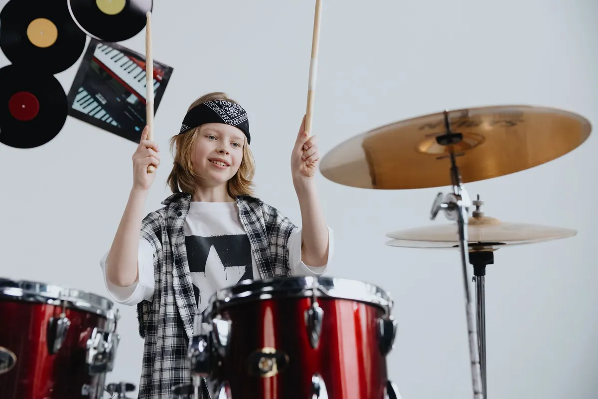 A grinning child at a drum kit with both sticks raised