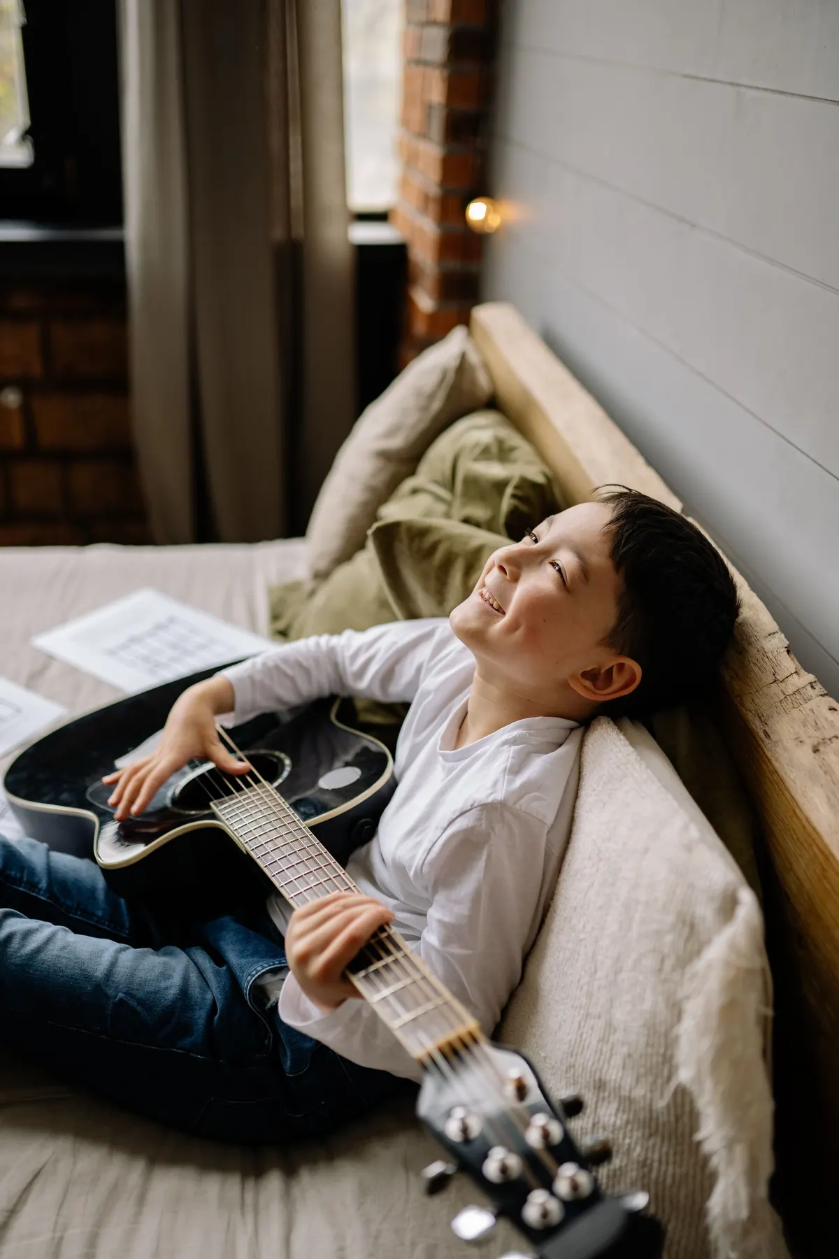 A smiling boy playing an acoustic guitar on his bed